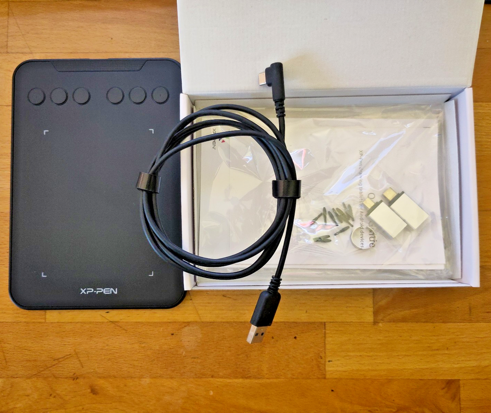 Links das Zeichentablett in schwarz mit Knöpfen und Bereich zum Zeichnen, daneben der Karton mit USB-A auf USB-C Adaptern, Ersatzstifteinsätzen und USB-C zu USB-A-Kabel.